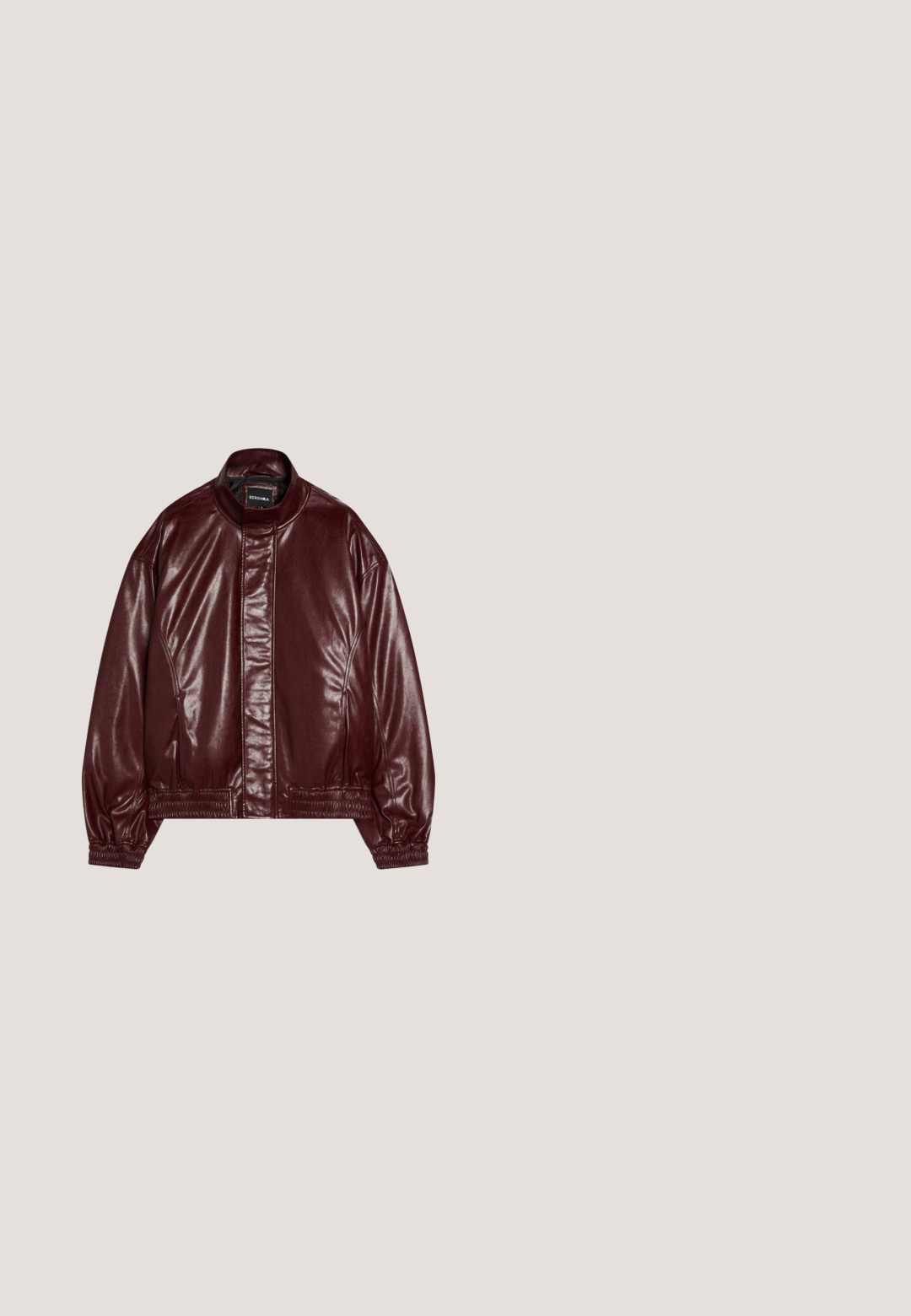 Bordeauxfarbene Kunstlederjacke mit Stehkragen, vorderem Reißverschluss unter einer Klappe, elastischen Bündchen und Saum, mit "Bershka"-Etikett.; Schwarzes, lässiges Baumwoll-T-Shirt mit weißem, abgenutztem Text: "ATELIER RETERNITY 2018 KREATIVDIREKTOR" zentriert auf der Vorderseite. Kurze Ärmel.; Dunkelgrüne Jeans aus Denim mit geradem Beinschnitt, fünf Taschen, Kupferfarbene Beschläge und einem subtilen Waschmuster.; Schwarze wasserdichte Gummistiefel mit strukturiertem Finish, ausgestattet mit einem flexiblen Schaft, einem Schnallenriemen und einer robusten, rutschfesten Sohle.; Schwarze halbrandlose Sonnenbrille mit rechteckigen dunklen Gläsern und schlanken, gebogenen Bügeln, die auf einer weißen Oberfläche liegen.; Schwarzes strukturiertes Leder rechteckige Tasche mit einem seitlichen Schnallenriemen und einem kleinen metallischen dreieckigen Logo in der vorderen Mitte.