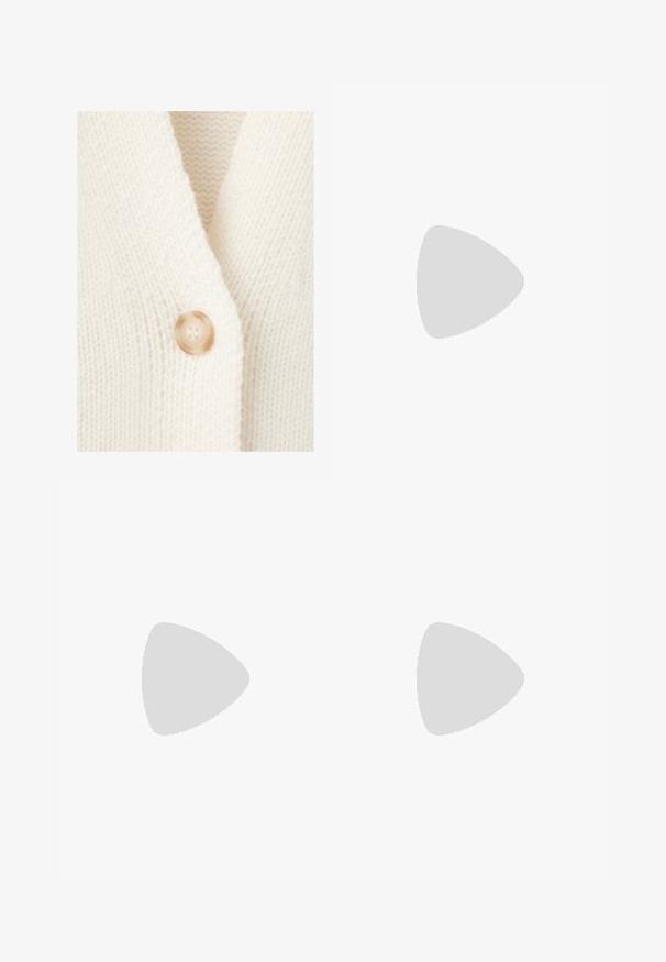 Gros plan d'un tissu tricoté couleur crème avec un seul bouton rond marron clair cousu sur le bord.; Haut court blanc sans manches à encolure ronde avec épaules larges et étiquette noire "B6" à l'intérieur du col.; Pantalon beige à jambes larges avec taille élastique et cordon, en tissu doux texturé.; Baskets Adidas Spezial blanches avec des rayures noires et une semelle en gomme flottant au-dessus de marches en pierre grises à l'extérieur.