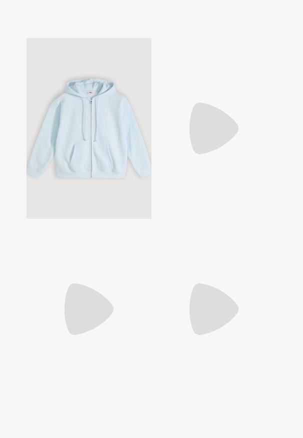 Lyseblå hættetrøje med lynlås lavet af blødt stof, med en forlommer foran, snøre i hætten og diskret logodetalje på brystet.; Hvid kortærmet t-shirt lavet af letvægts bomuld. Har en rund halsudskæring og rullede ærmer. Rød logoetiket på kraven.; Sorte bukser med høj talje og vidde forneden med små sideslids ved bunden og et lilla mærke i taljen.; Person iført hvide høje Converse-sneakers med tykke såler og beskårne blå jeans med flossede kanter mod en murstensvæg og hvidt gulv.