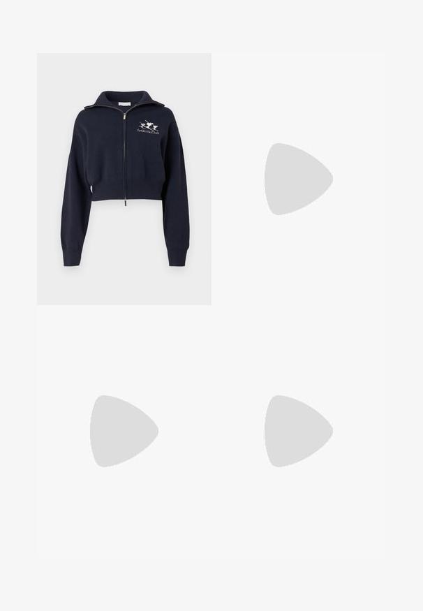 Sweat-shirt bleu marine court à zip avec col montant, doté de poignets et d'une ceinture côtelés, avec un logo blanc brodé sur le côté gauche de la poitrine.; Débardeur blanc à côtes avec un col rond, présentant le logo noir "KARL LAGERFELD JEANS" à l'avant.; Jupe beige longueur genou avec passants de ceinture, fermeture à boutons sur le devant, deux plis avant et coutures visibles sur fond uni.; Ballerines noires en cuir avec un bout pointu, une semelle plate et une petite sangle avec un détail de boucle en argent.