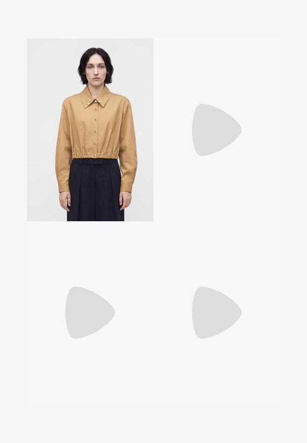 Mujer con cabello corto y oscuro que lleva una camisa corta de color marrón claro abotonada y pantalones anchos de cintura alta de color oscuro, de pie frente a un fondo liso.; Suéter beige de manga larga con cuello en V, puños y dobladillo acanalados, tejido liso, presentado sobre un fondo blanco.; Vaqueros de mezclilla anchos de color azul claro con una cintura alta, cierre de botón y cremallera, y bolsillos frontales.; Bailarinas de terciopelo burdeos con punta redondeada y tacón bajo, de textura suave y diseño elegante.