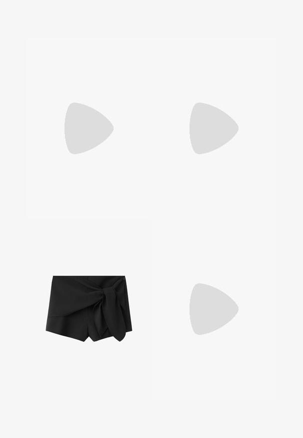 Haut noir sans manches à col halter, avec boutons sur le devant et deux petites poches devant, noué au cou avec un nœud.; Short noir taille haute avec détail en tissu superposé et grand nœud noué sur le côté droit devant.; Jambes portant des leggings côtelés foncés associés à des ballerines noires avec de petits détails en nœud sur fond blanc.; Sac hobo en cuir noir au design décontracté, avec une large bandoulière et une texture lisse. Dispose d'une fermeture éclair unique sur le côté.