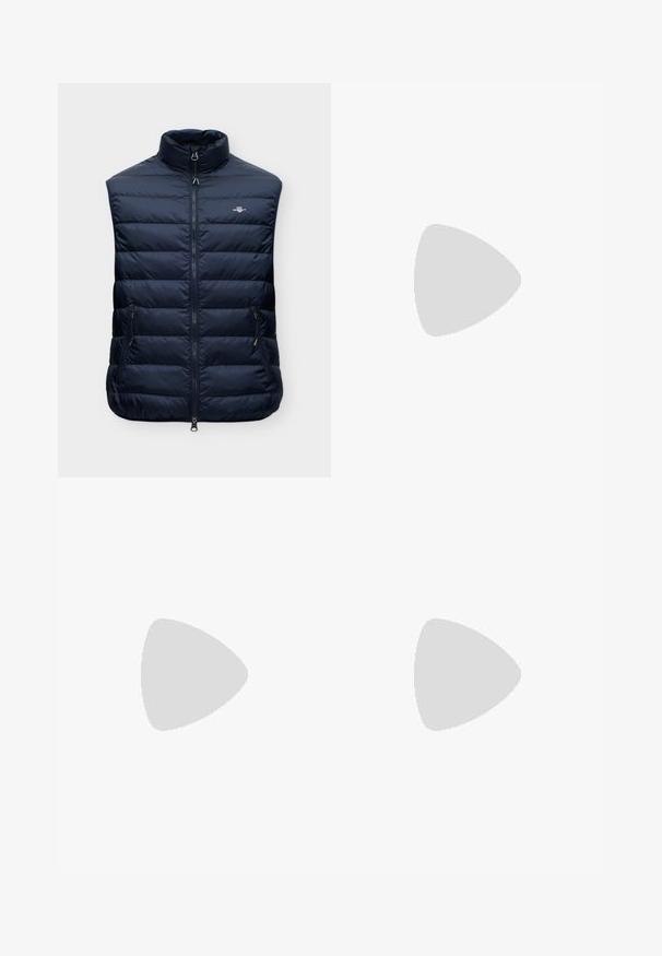 Veste sans manches matelassée bleu marine avec col montant, fermeture éclair à l'avant, deux poches latérales zippées et petit logo sur la poitrine.; Chemise bleu à manches longues avec un petit motif de lettres blanches, col italien et boutons noirs sur le devant.; Pantalons en laine bleu marine avec une coupe ajustée, présentant un devant plissé et des jambes larges. Texture lisse et lignes épurées, adaptés aux occasions formelles.; Mocassin en cuir noir brillant avec un détail de bride, posé sur un tapis beige près d'une table basse ronde en bois brun dans une pièce doucement éclairée.
