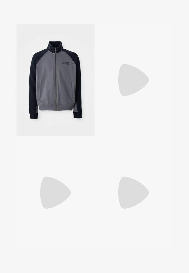 Harmaa ja musta vetoketjullinen takki korkean kauluksen kanssa, jossa on kaksi etutaskua ja "Dickies Workwear Company" -logo rinnassa.; Valkoinen puuvillapaita lyhyillä hihoilla, pyöreä kaula-aukko, jossa on pieni kultainen logo vasemmassa rintapielessä. Pehmeä kangas ja normaali istuvuus.; Mustat, leveälahkeiset farkut, joissa on etunapitus, vyölenkit ja kaksi sivutaskua, joissa on sileä pinta.; Mustat ja valkoiset Nike Air Force 1 -tennarit, joissa on nahkavarsi, rei'itetty varvas ja teksturoitu pohja, käytetty vaaleanbeigejen käännettyjen housujen ja valkoisten sukkien kanssa.