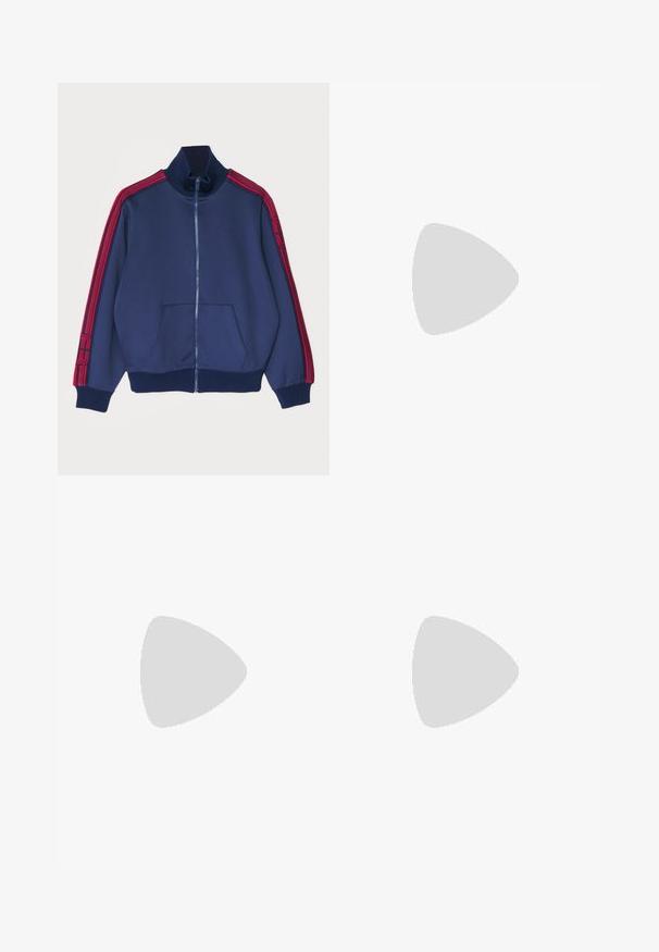 Giacca a track in tinta blu navy con zip e colletto alto, dettagli a strisce rosse sulle maniche e tasche frontali. Tessuto morbido con polsini a costine.; Abito a maglia rosso a coste con maniche lunghe, corpino aderente e gonna svasata. Presenta un collo rotondo e cinque bottoni bianchi davanti.; Mocassino in pelle verniciata nera con finitura liscia, dettagli cuciti nella parte anteriore e tacco basso per un supporto ottimale. Accentatura dorata sulla fascia.; Borsa a tracolla in camoscio grigio con frange, grande apertura con manico superiore e una tracolla sottile. Il modello indossa una giacca blu navy e una gonna rossa.