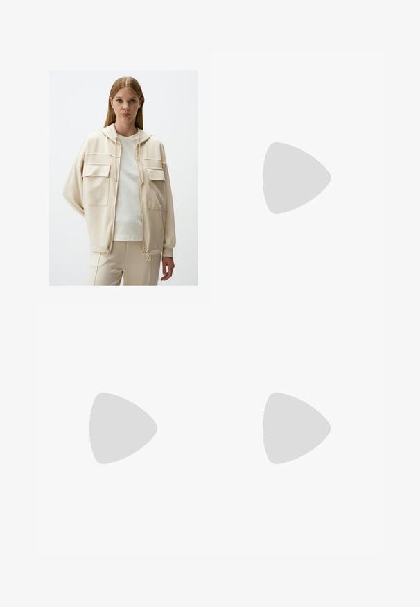 Beige gewatteerde jas met twee voorzakken, trekkoord aan de onderkant en zachte stof, gedragen over een lichte blouse en bijpassende broek.; Zwarte top met één schouder, gemaakt van zachte, rekbare stof met een aansluitend ontwerp en geplooide details aan de zijkant. Inclusief dunne verstelbare band.; Zwarte, op maat gemaakte broeken met een gladde afwerking, recht gesneden ontwerp en een tailleband. Beschikt over subtiele stikdetails en heeft geen zakken.; Zwarte ballerina's van soepel leer met een ronde neus en een lage, platte zool, met een slip-on ontwerp en zonder zichtbare hardware.; Zwarte, hoekige zonnebril met grijze lenzen; gemaakt van plastic, met dikke armen met een gladde oppervlakte en zonder zichtbare hardware.