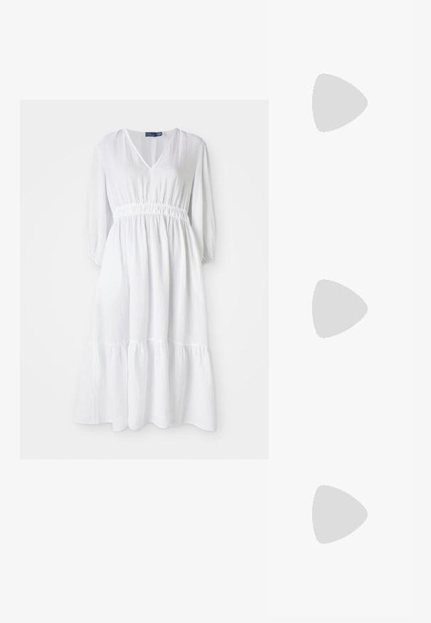 Witte jurk met V-vormige halslijn, elastische taille en driekwartmouwen. Heeft een gelaagd ruches uiterlijk aan de onderkant en een gestructureerde stof.; Witte leren sneakers, laag model, met een gladde textuur, ronde neus, vetersluiting aan de voorkant en een klein geborduurd logo aan de zijkant.; Zwarte leren halfronde tas met een gouden hardware accent, voorzien van een ritssluiting. Draagt crossbody over een grijze trui.; Gouden knoopstuds met een gedraaide design. Elke oorring heeft een ronde transparante achterkant voor een veilige draagervaring.
