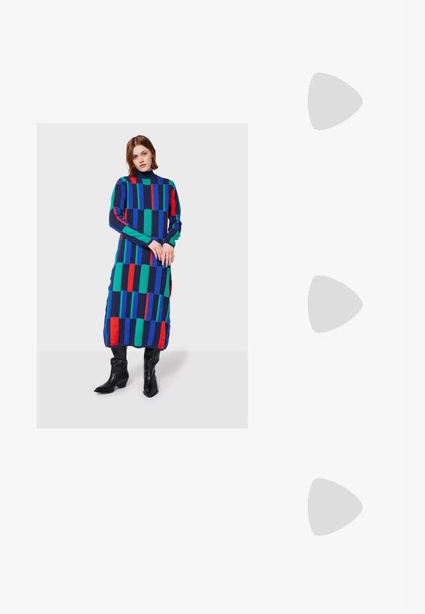 Kobieta stoi w długiej, kolorowej sukience w geometryczny wzór z niebieskimi, zielonymi i czerwonymi paskami, w zestawieniu z czarnymi, wysokimi kowbojkami.; Bershka - Sandały na obcasie; Brązowe okulary w motyle z owalnymi soczewkami, cienkimi ramionami i przezroczystymi akcentami. Posiadają subtelne logo na zauszniku. Smukły, lekki design.; Brązowa skórzana torba na ramię o zaokrąglonym kształcie, ze złotymi akcentami i zapięciem w formie klamry. Posiada odpinany pasek na ramię.