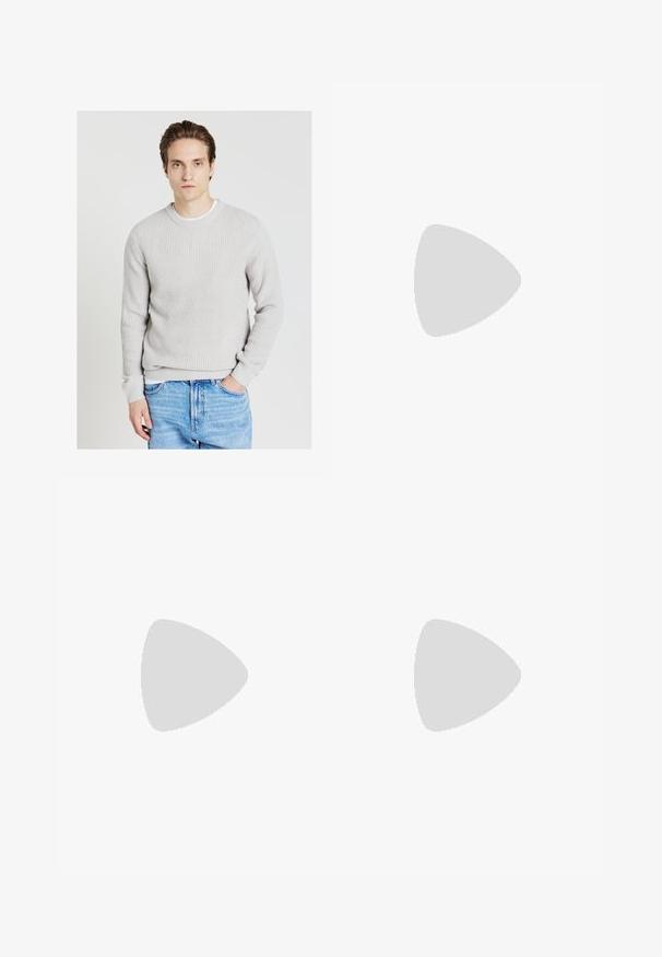 Junger Mann mit hellbraunem Haar, der einen hellgrauen Strickpullover und eine blaue Jeans trägt, steht mit einer Hand in der Tasche vor einem weißen Hintergrund.; Schlanke Jeans aus ausgebleichtem grauem Denim, mit abgenutzten Details an den Knien, kombiniert mit weißen Sneakers. Glatte Textur, schlanker Schnitt.; Weiße Leder-Sneaker mit Perforationen, runder Zehenpartie, flacher Sohle und dicken Schnürsenkeln, mit sichtbar eingesticktem Swoosh-Logo und "AIR" an der Ferse.; Silberner Armbanduhr aus Edelstahl mit einem dunkelblauen Zifferblatt, metallischen Stundenmarkierungen und einem Datumsfenster bei 3 Uhr. Wasserdicht bis 50 Meter.