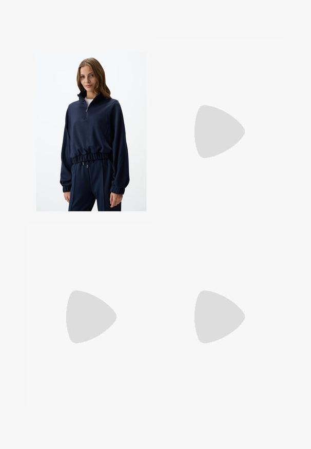 Pullover navy a mezza zip in tessuto morbido, con polsini ed orlo elastici, progettato con una vestibilità rilassata e sottili dettagli in oro.; Pantaloni larghi beige realizzati in cotone morbido, dotati di una cintura e tasche laterali. Il tessuto presenta una texture liscia con risvolti piegati.; Sandalo in pelle marrone con fasce incrociate, suola piatta e una texture liscia. Design semplice senza decorazioni o motivi.; Borsa in pelle marrone con parte superiore arricciata e tracolla a catena. Texture liscia con finitura lucida e dettagli di cucitura sottili sui lati.