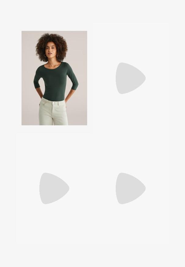 Frau mit lockigem Haar, die ein grünes Oberteil mit drei Viertelärmeln und beigefarbene Hose trägt, steht mit den Händen hinter dem Rücken vor einem schlichten Hintergrund.; Tory Burch TWILL SKIRT - Maxirock - blue abstract; Weißer Sneaker mit Obermaterial aus Mesh und Wildleder, runder Zehenpartie und gepolstertem Kragen. Verfügt über reflektierende Akzente und eine dicke, strukturierte Sohle.; Braune Leder-Crossbody-Tasche mit Logo-Muster, goldener Kettenriemen, Reißverschlussverschluss und goldenen Hardware-Details. Kompakte und rechteckige Form.